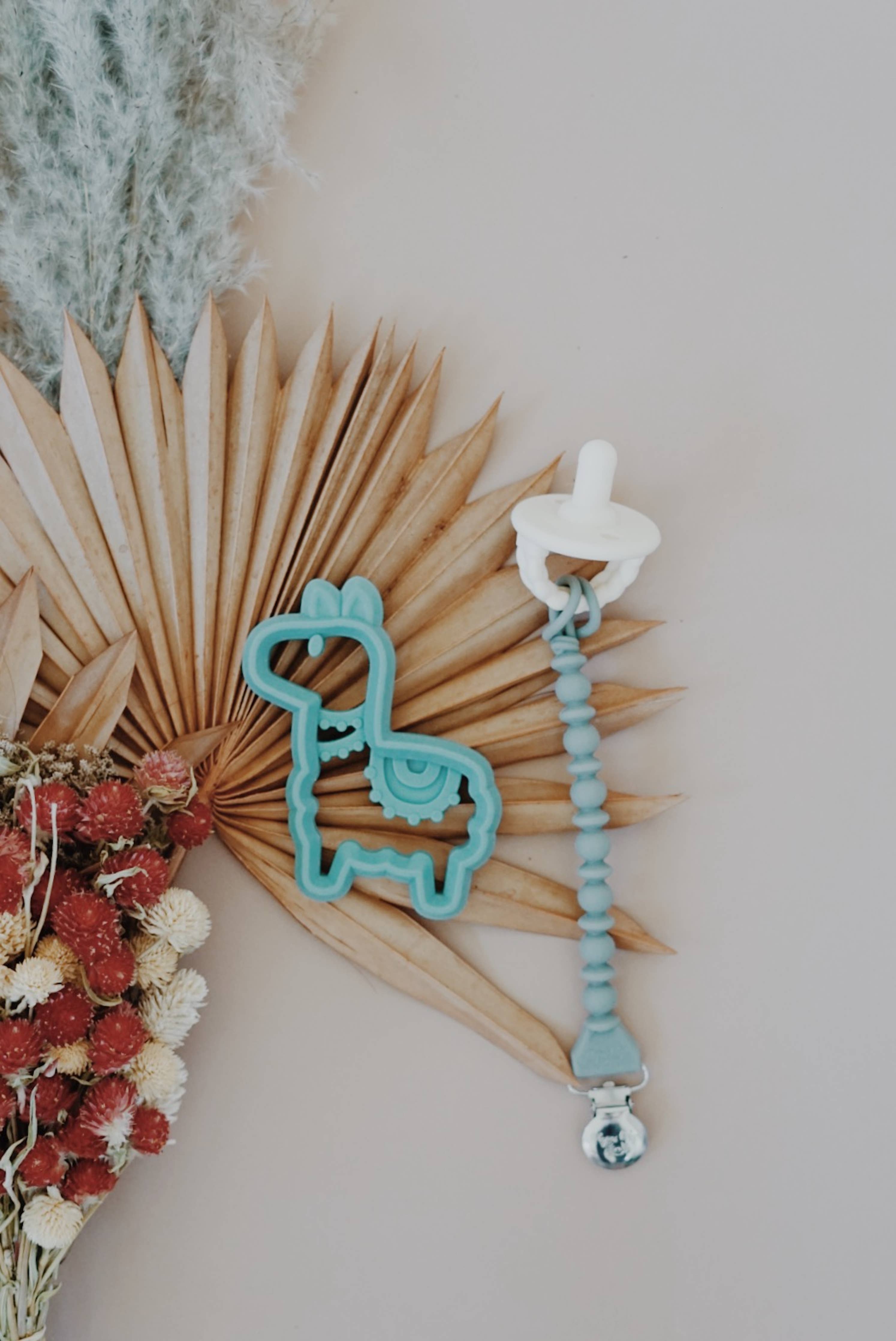 Sweetie Strap™ silicone pacifier clip with llama design, stylishly holding a pacifier against a backdrop of dried flowers.