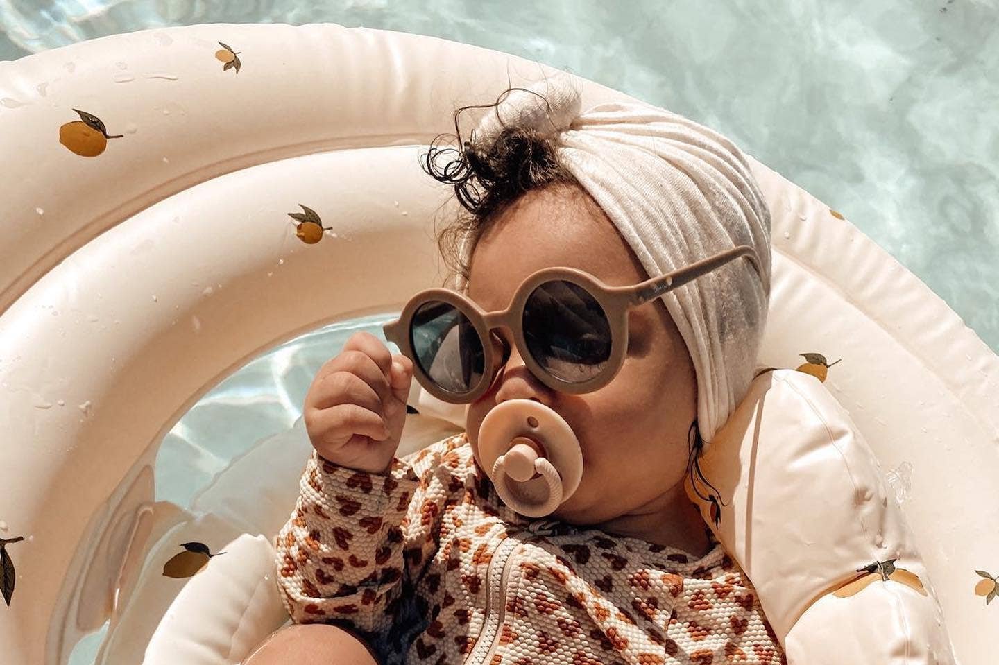 Cute baby in sunglasses enjoying the pool, relaxing with an Itzy Sootherβ’ Natural Rubber Pacifier.