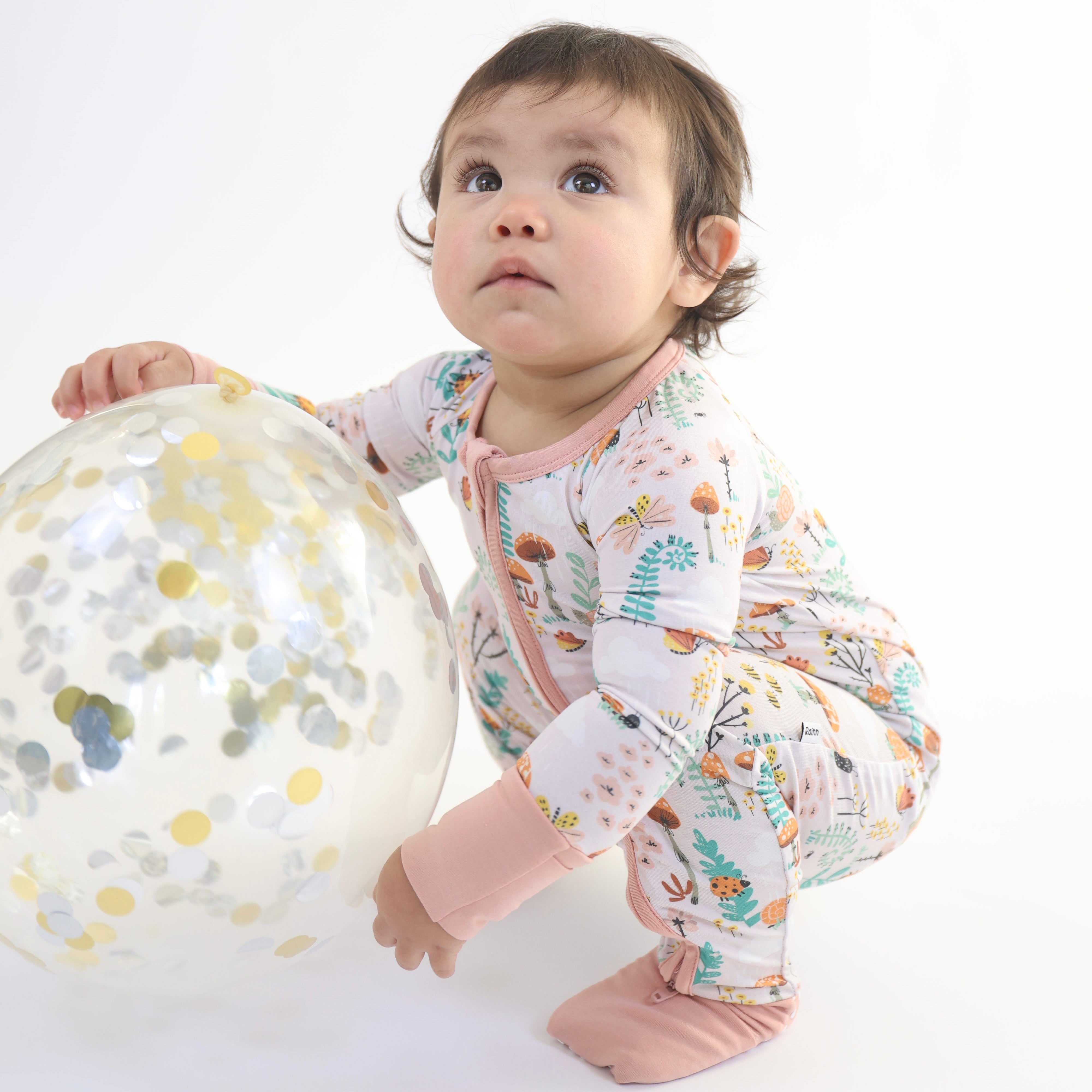 Adorable baby in floral patterned pajamas playing with a clear balloon filled with colorful confetti.