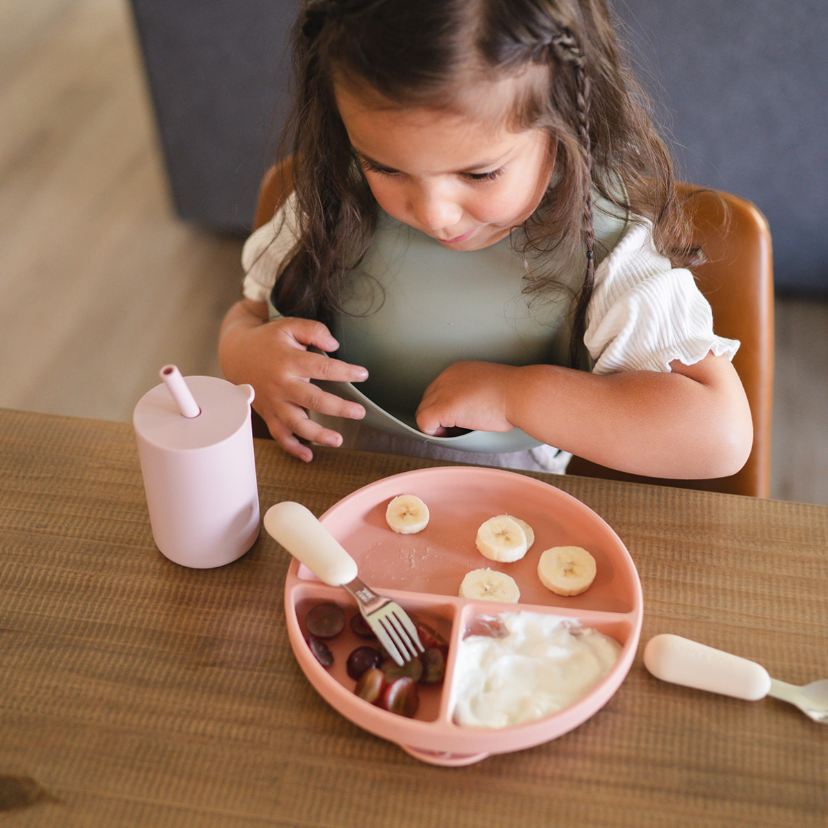 Cute girl enjoying a healthy meal of bananas, grapes, and yogurt on a colorful feeding plate.